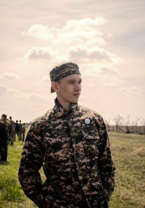 Portrait of a young soldier in camouflage uniform standing outdoors under a bright sky.