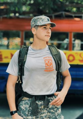 Young soldier in military uniform and cap poses outdoors. Bright day in a casual setting.